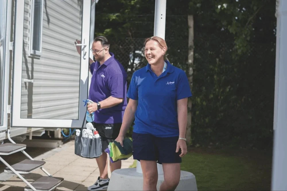 Two holiday park staff walking between caravans on a summer morning, cleaning kit in hand