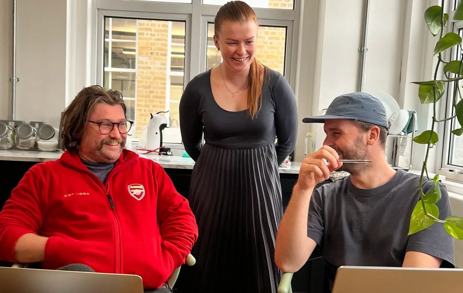 Three colleagues laughing over laptops at the office kitchen table