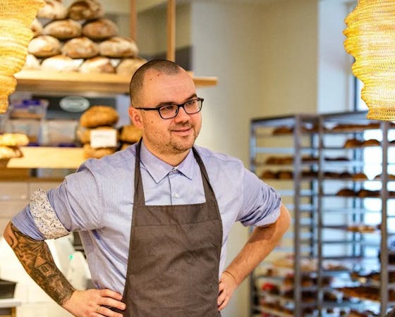 A smiling man with glasses stands in a bakery, wearing a blue shirt and apron. Shelves filled with bread are visible in the background, creating a warm, cozy atmosphere.