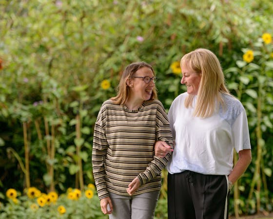 Image of two women walking side by side, engaged in conversation and making eye contact. The background is a blurred green landscape with sunflowers.