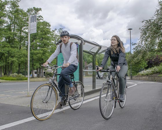 A man and a woman cycling past a bus stop with trees in the background.