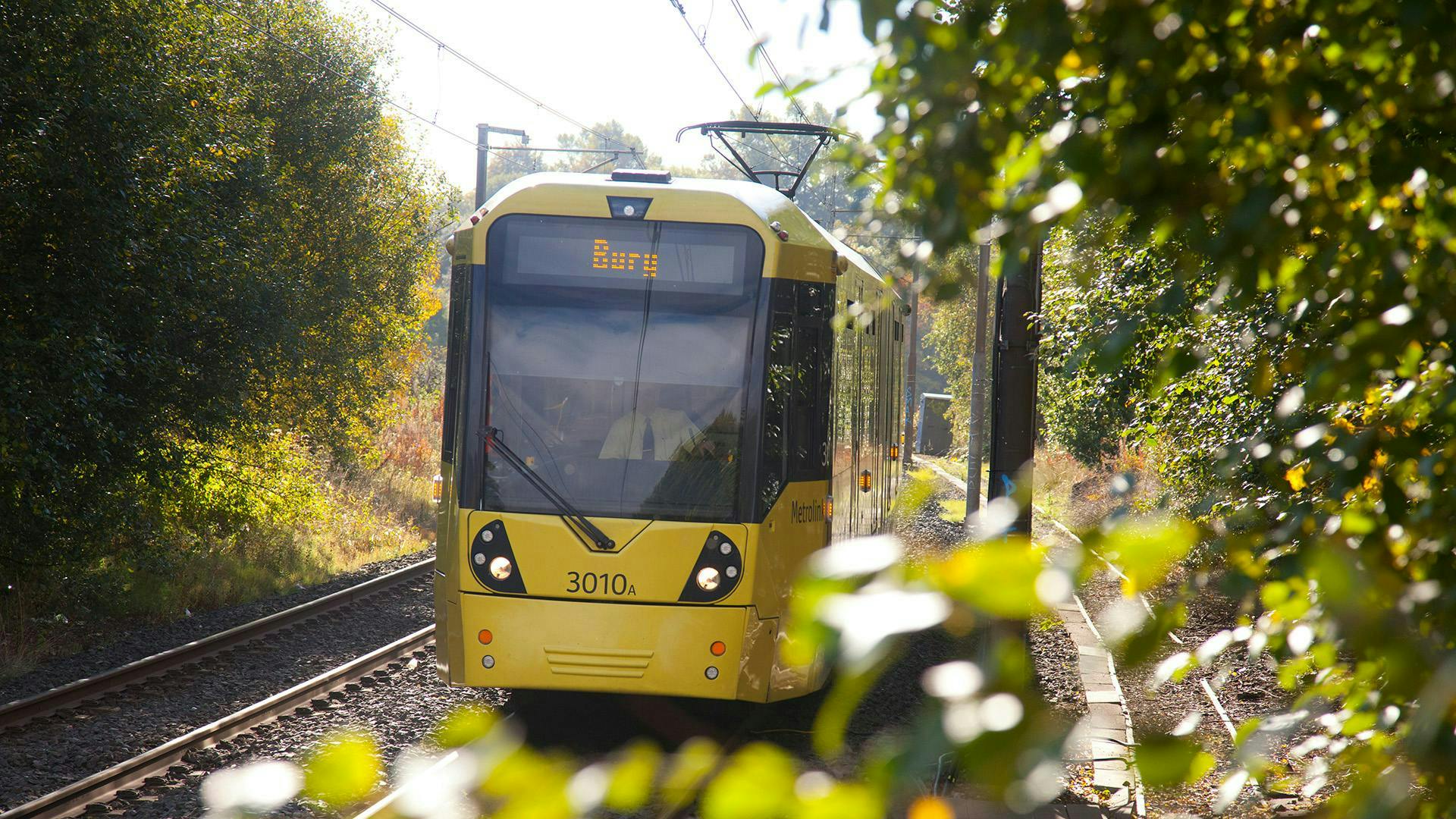 A yellow train with Bury as the destination, with some greenery at the forefront of the image.
