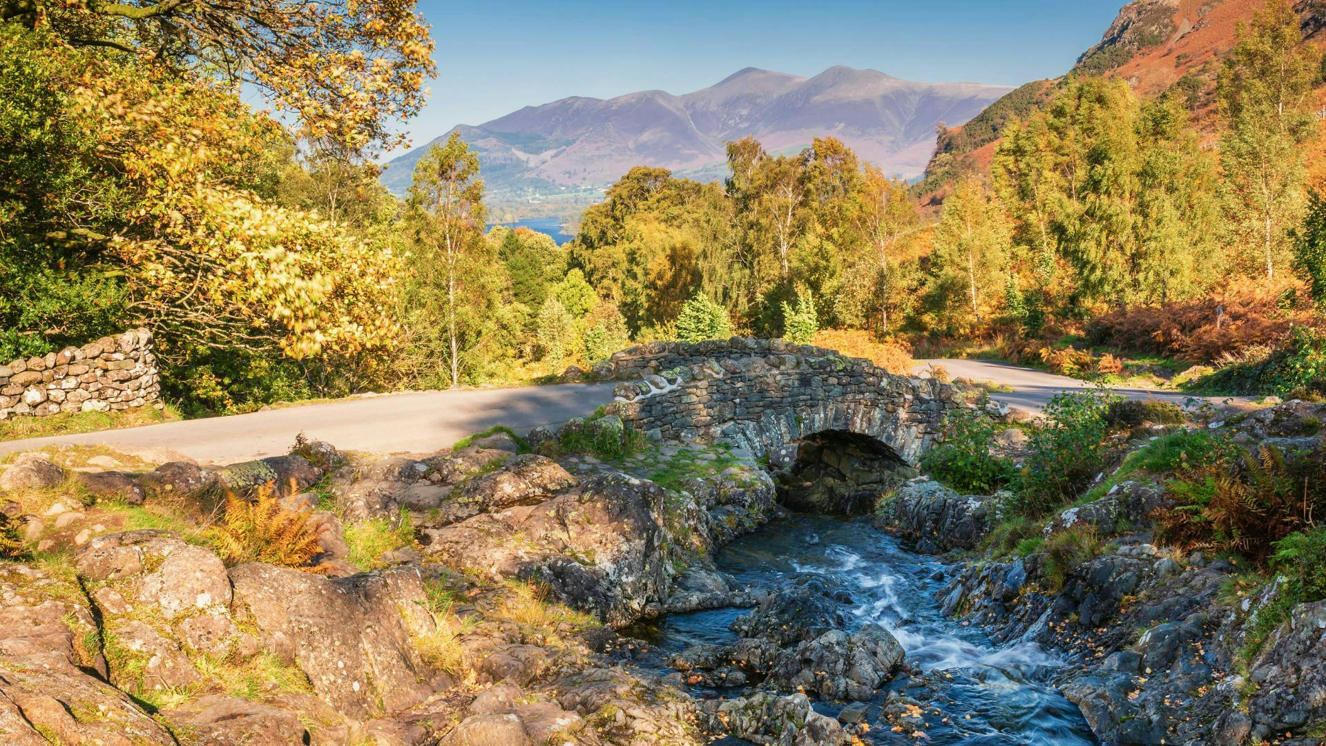 a old bridge with a small river flowing underneath with the peaks of mountains in the background