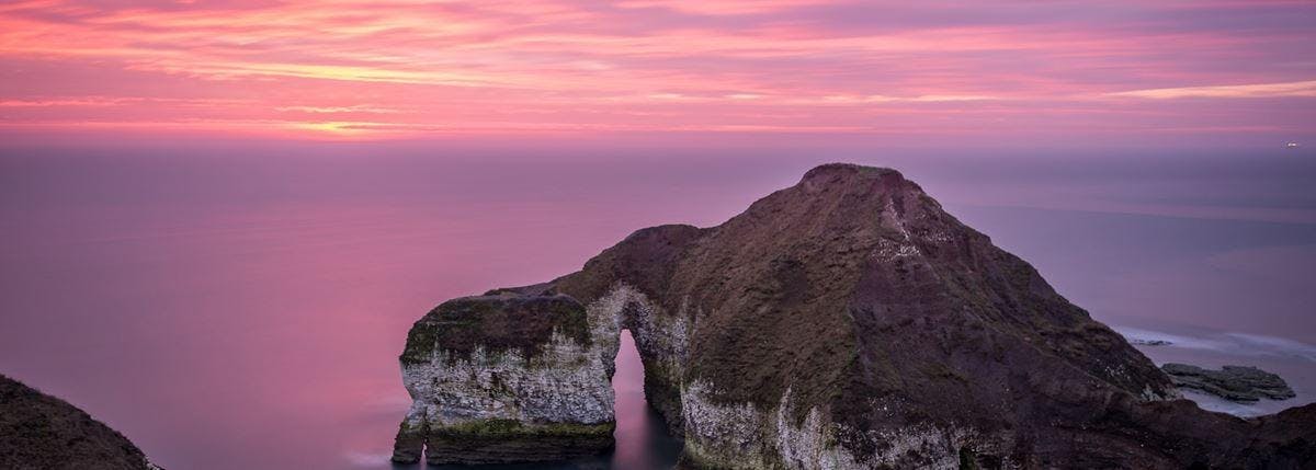 Image of a stunning pink sunset seen from a cliff, with another cliff in the foreground.