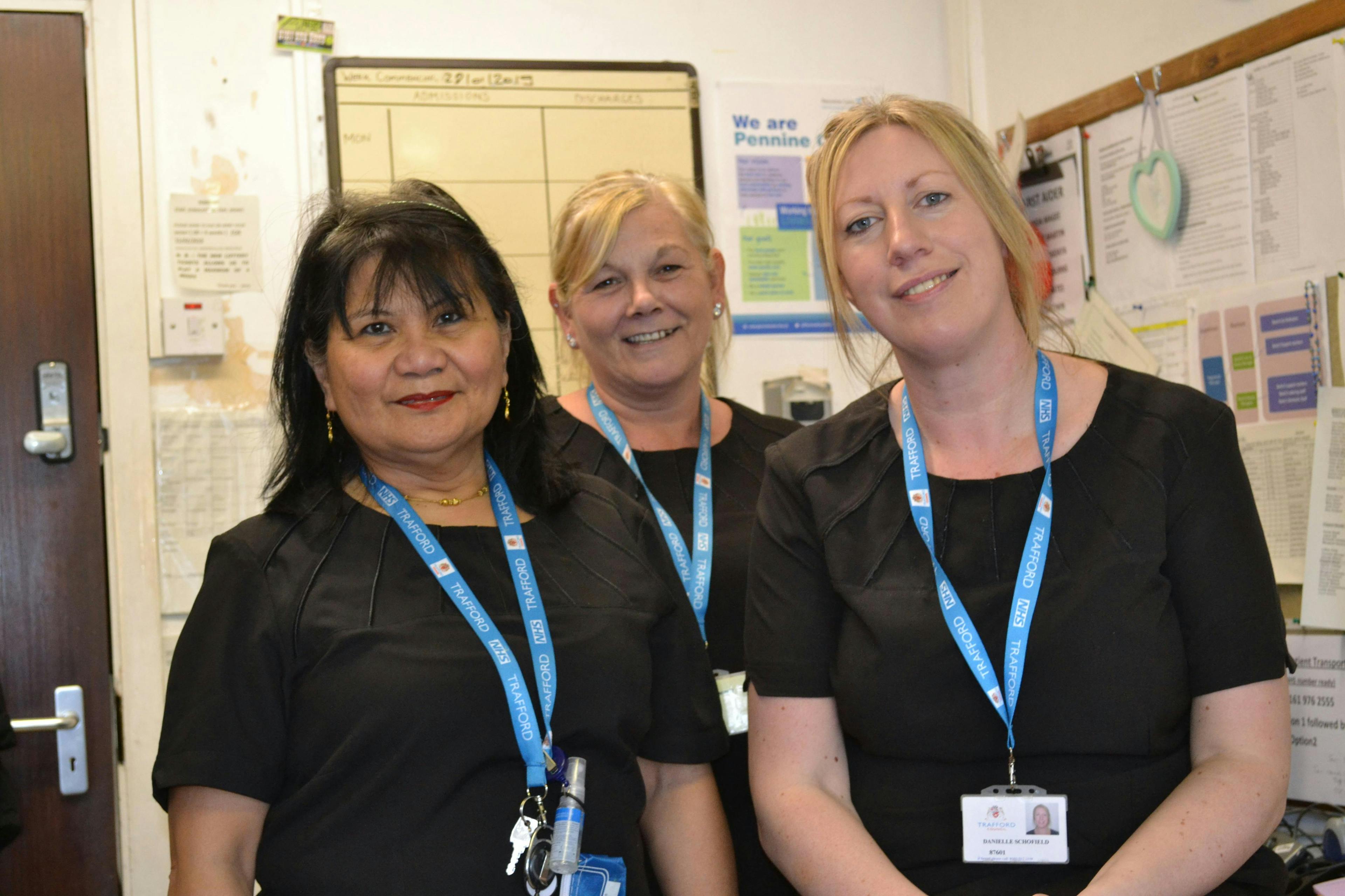 Three smiling ladies standing in an office, visible from the bust up. They wear blue ID badges. Behind them, a board on the wall holds papers, with a door in the background on the left side.