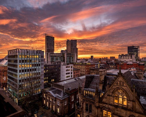A scenic view of Manchester city featuring modern high-rise buildings, historic architecture, and lush green trees under a sunset