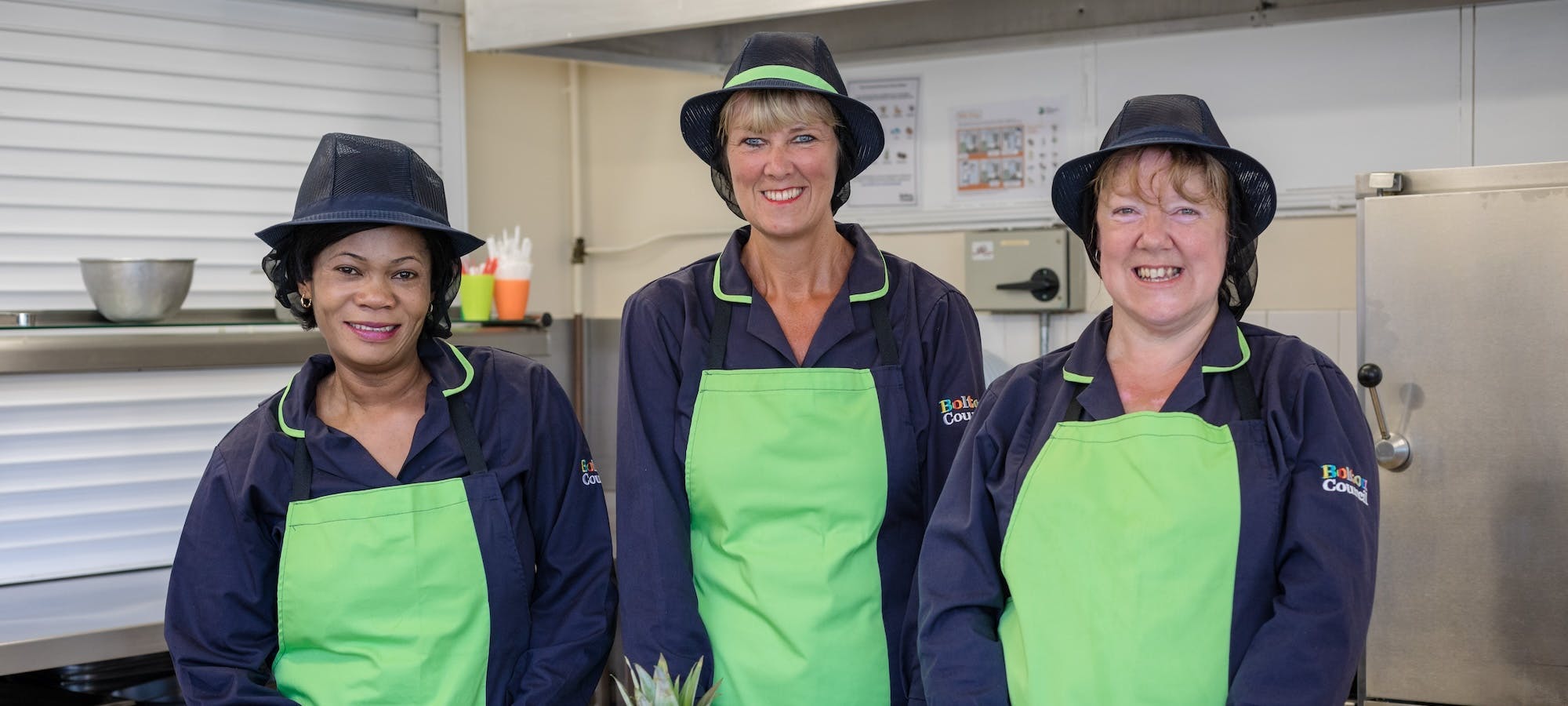 Image of three smiling chefs wearing green aprons in a kichen.
