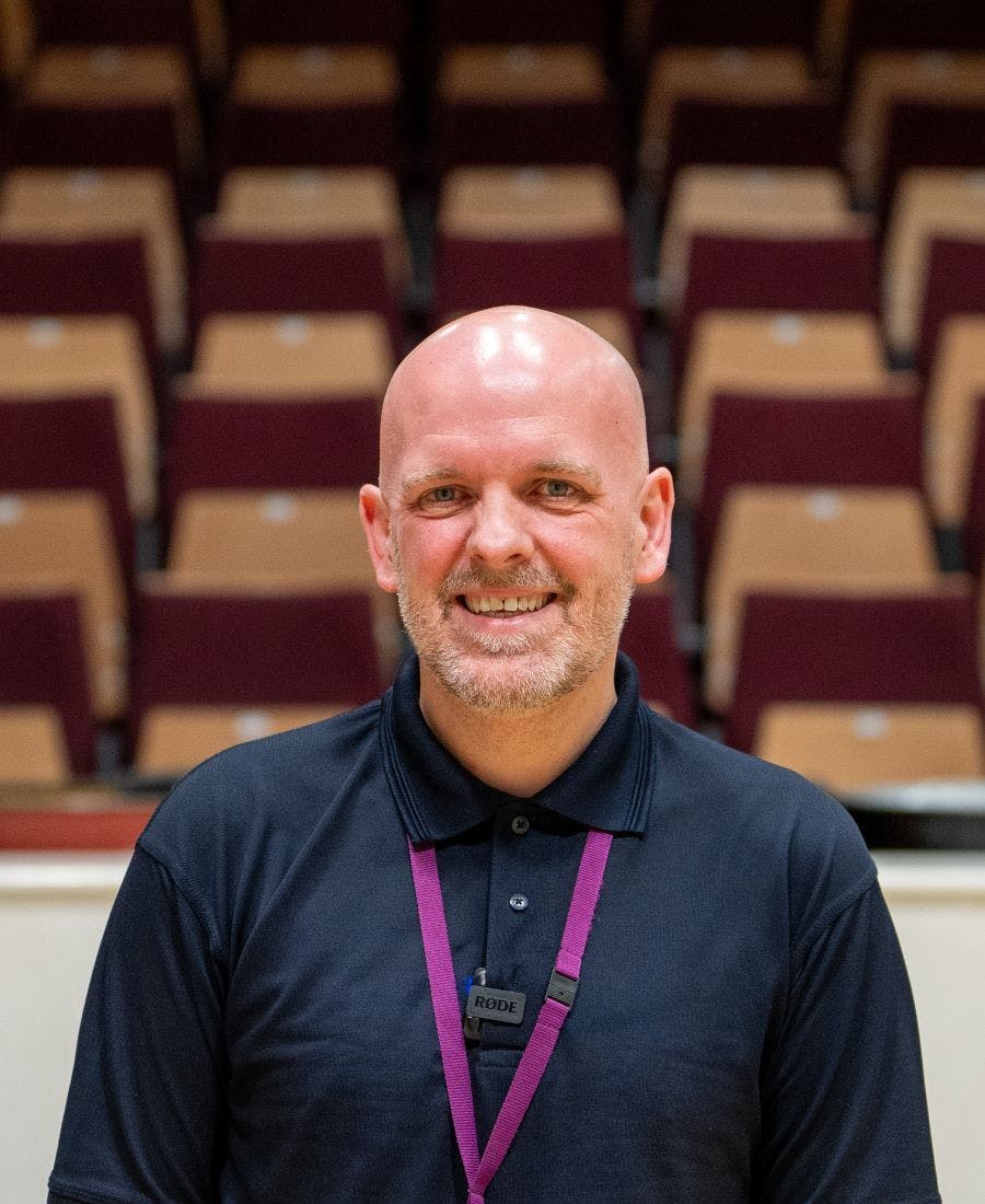 A man with a beard, wearing a dark polo shirt and a lanyard, smiles while standing in front of rows of empty seats in an auditorium.