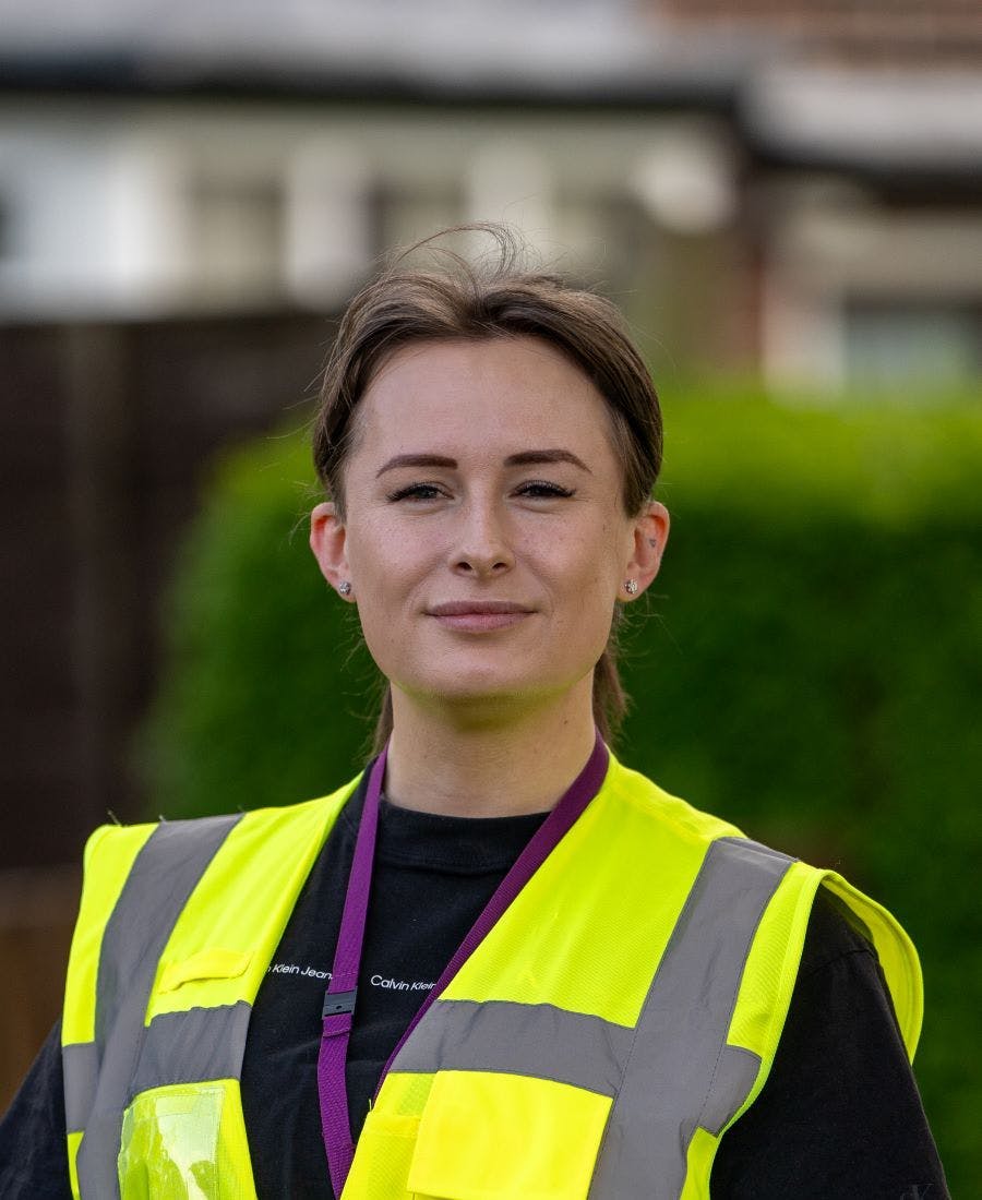 Portrait of a woman wearing a high-visibility safety vest and a lanyard, standing outdoors with a confident expression and greenery in the blurred background.