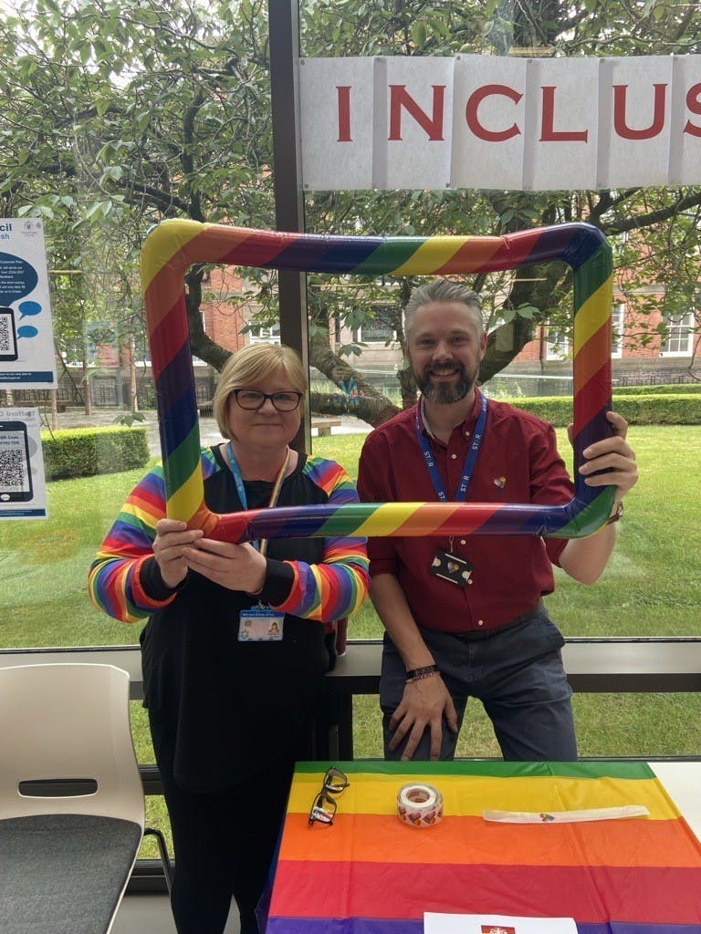 A smiling man and woman stand in front of a table with a pride flag, holding a colourful pride frame with their faces in it. In the background, there's an inclusive flag displayed.