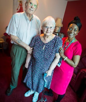 Image of three people in a room: an elderly couple and a young lady holding the elderly lady who has a cane to help her walk.